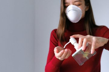 pexels-photo-3987146-3987146 A woman in a mask applies hand sanitizer for hygiene indoors during pandemic.
